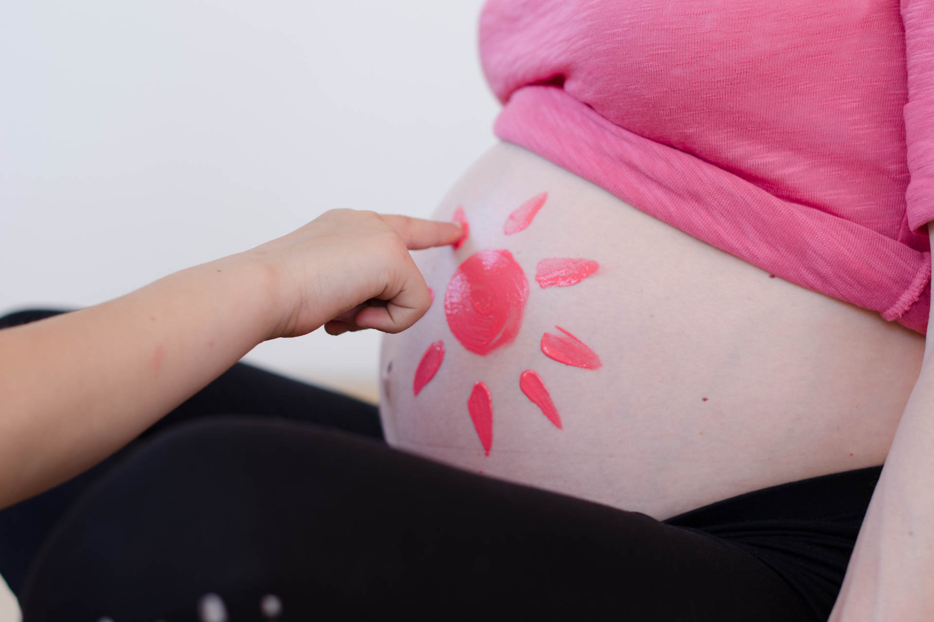 Bauch einer schwangeren Frau und eine Kinderhand die eine pinke Sonne auf den Bauch malt